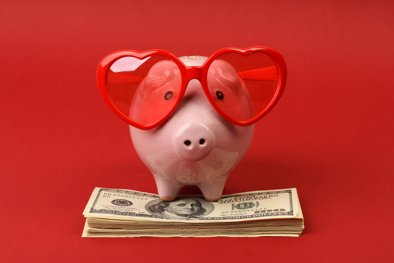 Piggy bank in love with red heart sunglasses standing on stack of money american hundred dollar bills on red background