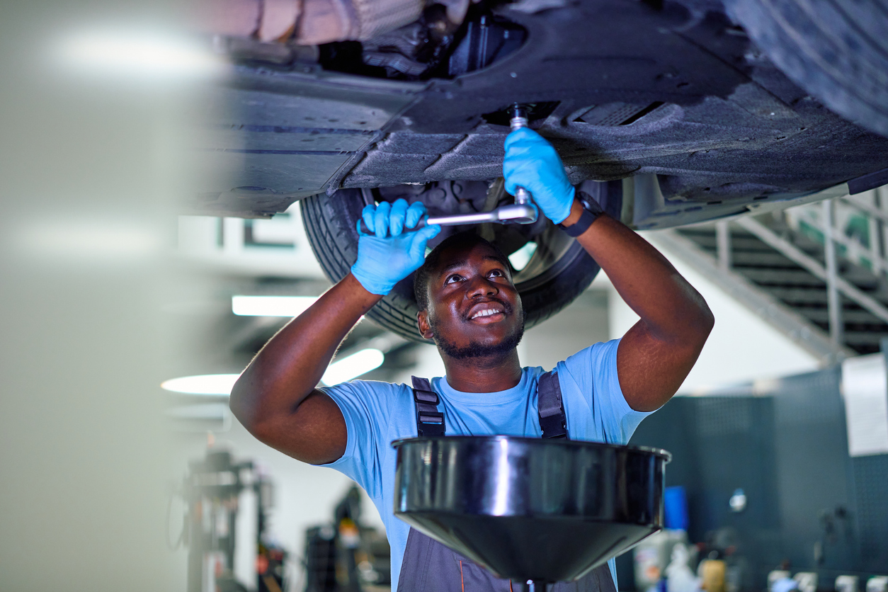 Smiling technician in blue gloves using a ratchet wrench to loosen a vehicle's oil drain plug, with a drain pan positioned below the car on a service lift.