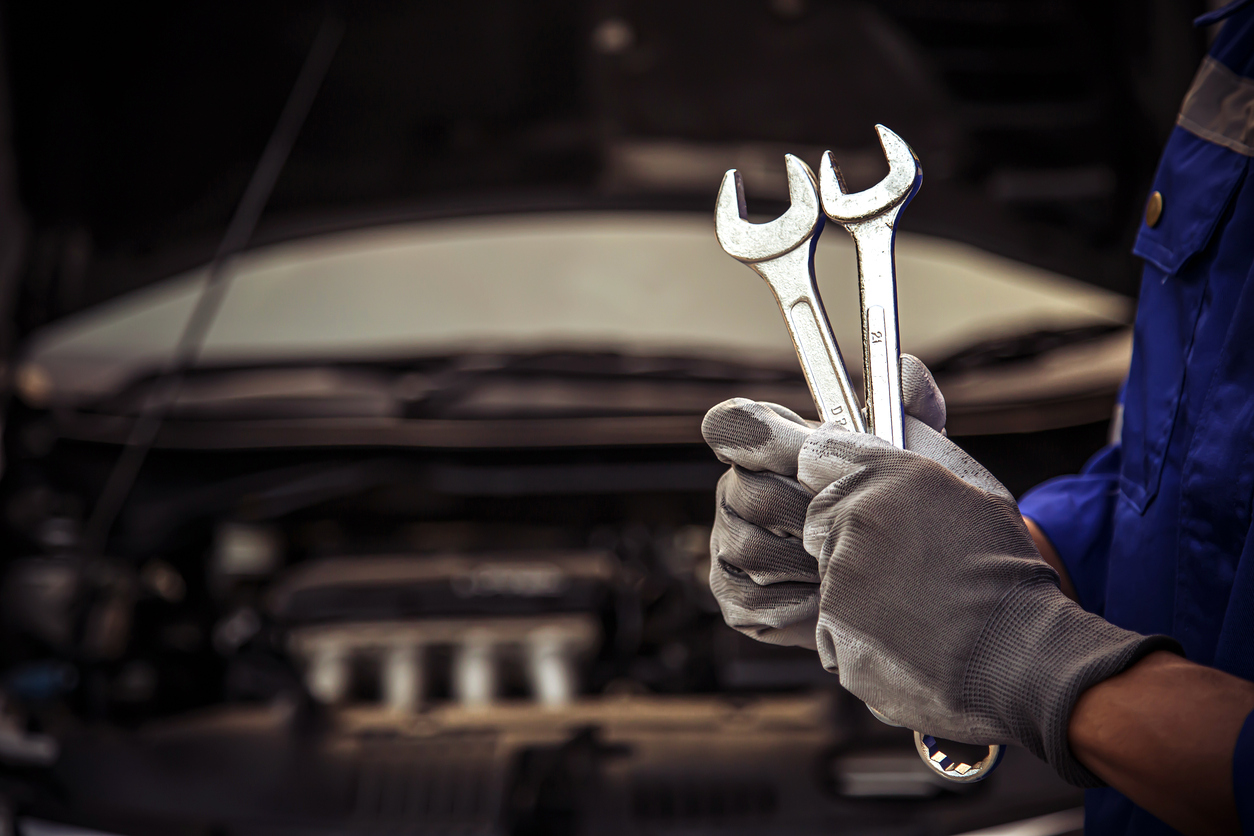 Gloved mechanic in a blue uniform holding two combination wrenches in front of a vehicle with its hood open.